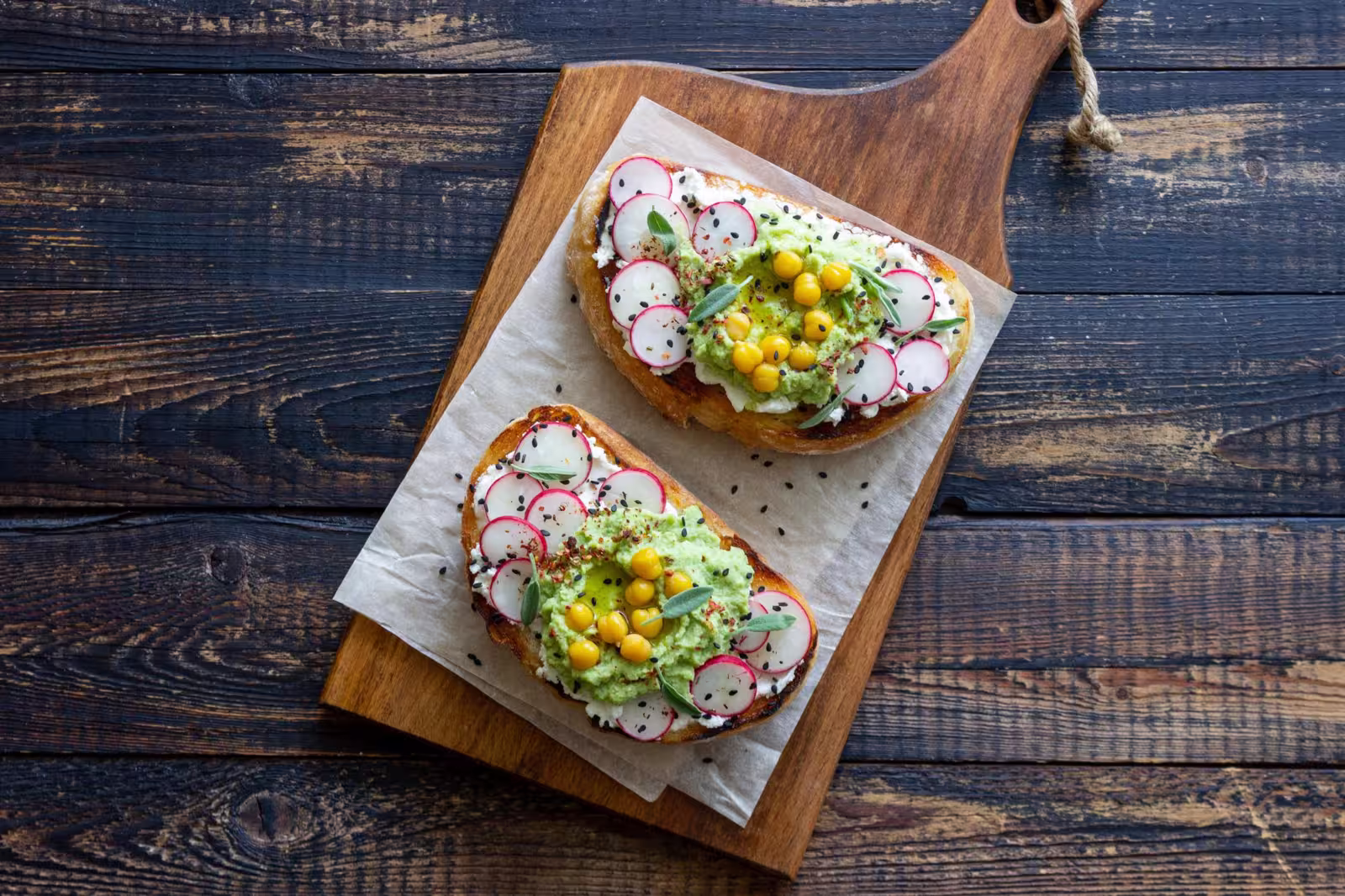 Slice of bruschetta topped with guacamole, radish slices, crumbled white cheese, chickpeas, and sage leaves