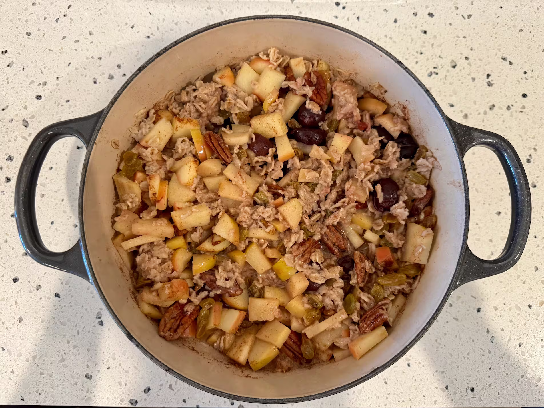 Downward view of oatmeal in a large gray pot. Apples, pecans, cherries, and oats are visible. Oats are clumped and dense